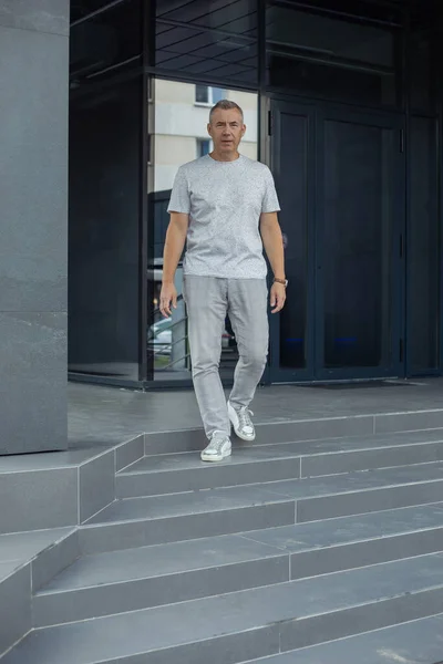 Vertical full length shot of confident gray haired man in gray outfit leaning on wall near entrance outdoors. Office worker, manager occupation. Recruitment, find new job and hr service in office