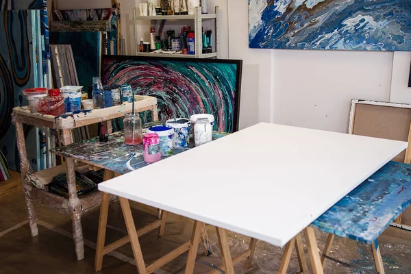 Interior of creative art workshop. Different buckets with paint, dirty glass jars, big white canvas on wooden desk stained, abstract picture. Modern art, creativity, decoration, workshop, painting.