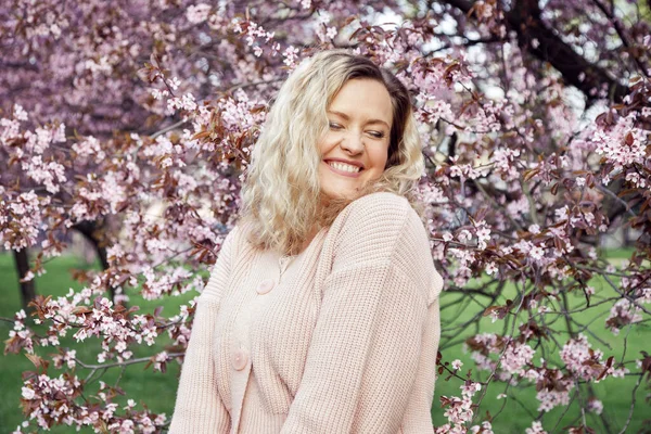 Portrait of young smiling wonderful woman with long wavy fair hair wearing pink knitted cardigan, standing near blossoming bird-cherry tree with white flowers, screwing up eyes in park. Spring.