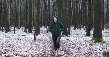 Happy middle aged woman in winter forest, go forward to camera on wet snow path in park. Adult woman in hood on head of green jacket from eco fur. Smiling person walk outdoors among bare trees.