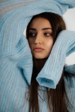 Portrait of young thoughtful woman with long dark hair, natural make-up wearing blue knitted dress, holding hands over head looking aside on white background. Beauty, fashion, vogue. Vertical, studio.