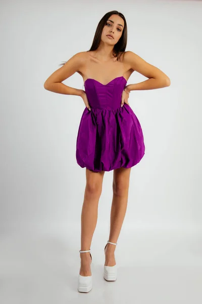 Portrait of young stunning woman with make-up wearing purple strapless balloon dress, high-heeled shoes, looking at camera, standing, holding hands on waist on white background. Beauty. Vertical.