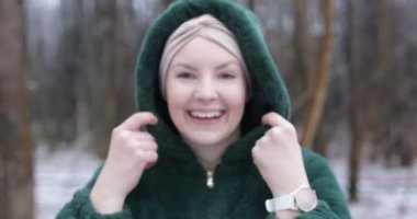 Laughing middle aged woman in winter forest closeup. Female portrait in snow park. Cheerful adult woman with headband in hood of green jacket from eco fur. Happy woman walk outdoors among bare trees.