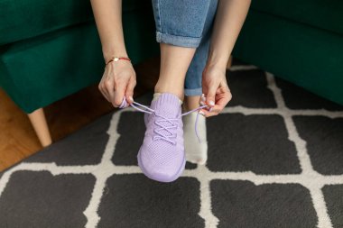 Cropped photo of woman wearing jeans, sitting on green sofa at home, lacing up trying on violet sneakers, stretching laces, getting ready for walking, running, sport, training workout. Sportswear.