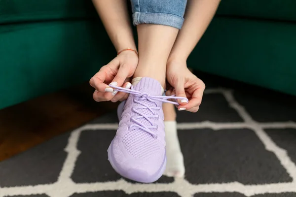 Female hands lace up violet running sneaker sitting on sofa at home room closeup. Shoes lacing, tying bow. Comfortable sneakers for walk, stylish textile footwear. Active lifestyle and sport.