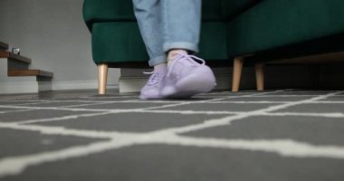 Unrecognizable woman wearing jeans, going along green sofa at home, trying on violet sneakers, raising heels, stretching legs, preparing for running, sport, training. Sportswear, active lifestyle.