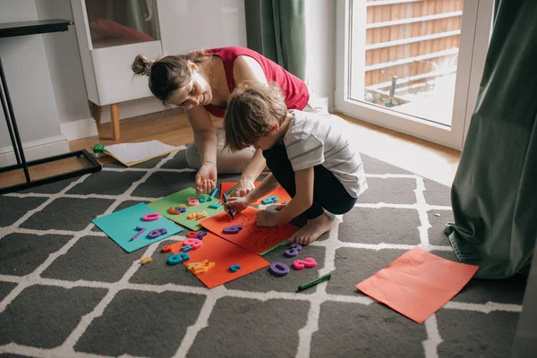 Happy mother and little son play together sitting on carpet at home room. Draw markers on colored paper sheets, learning numbers, numeration. Educational games, preschooler, home study.