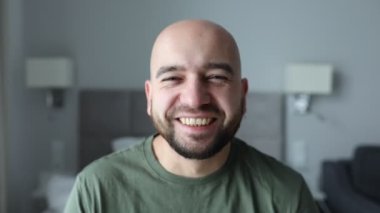Video of laughing, positive, relaxed bald unshaven man shaking head and looking at camera in gray bedroom. First day of weekend. Good mood after antidepressants, medical therapy result. 4k animation