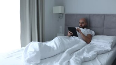 Mature bearded smiling man scrolling, typing on digital tablet screen, lying in white linen in bed in bedroom. Freelance work, trader occupation, remote work at home. Wireless technology. Video, 4k