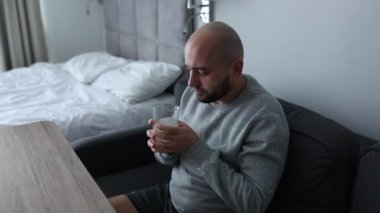 Tired mature bearded man drink hot water with dissolved medicine, hold glass with two hands, sit on sofa at home. Hangover after drunk condition. Home treatment. Removal of symptoms. Video full hd