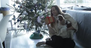 Beautiful happy woman with cute spaniel dog open christmas present sitting on floor under christmas tree. Unwrapping christmas gift at home, celebrating New Year. Festive home decoration.