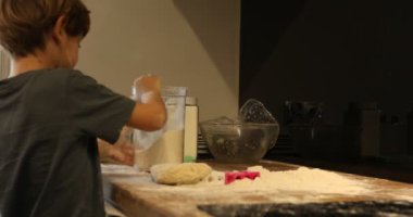 Side view of happy dirty preteen boy playing with flour dough, pouring flour on plastic toy from container on wooden table, baking in kitchen, having fun, making mess at home. Childhood, cooking.