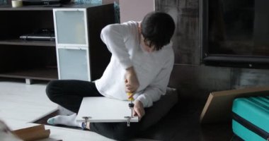 Portrait of busy teenage boy wearing white sweatshirt, black trousers, sitting on blanket on floor, tightening screw in hinge of door of bedside table with yellow screwdriver with hand, installation.