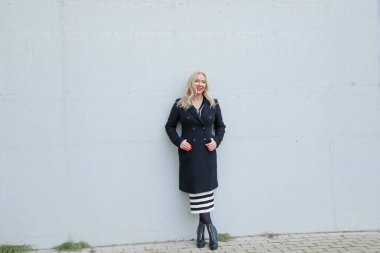 Successful blonde woman in elegant black coat with smile posing on gray building wall. Female full length photo. Stylish clothes, seasonal casual wear. Business people, outfit, autumn outerwear.