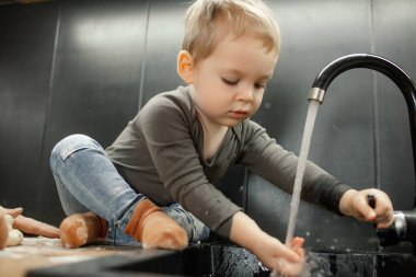 Portrait of little child washing hands in sink sitting on kitchen tabletop. Baby boy get dirty in scattered flour. Water tap, plumbing. Baking pastry, mess in kitchen. Family cooking.