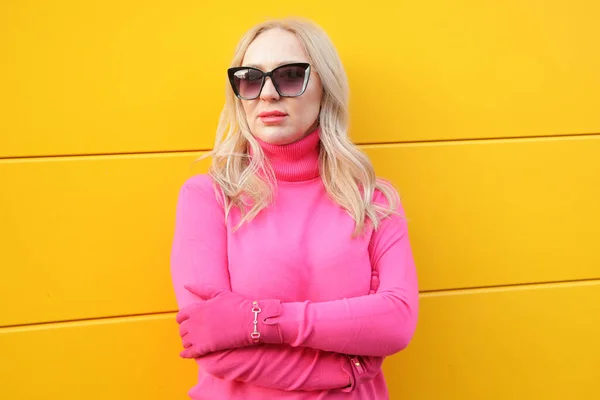 Successful blonde woman in bright pink turtleneck and gloves with sunglasses on yellow wall. Female portrait. Stylish clothes, seasonal trend color. Fashionable wear, colorful casual outfit.