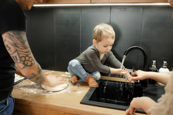 Portrait of little child washing hands in sink sitting on kitchen tabletop. Baby boy get dirty in flour. Dad knead dough with hands. Water tap, plumbing. Baking, mess in kitchen, water splashes.