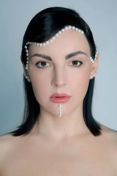 Graceful young woman with pearl beads on face. Female fashion portrait. Creative evening makeup with decorative details for event celebration. Woman with bare neckline. Modern beauty trend.