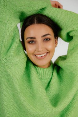 Close up vertical toothy smiling dark haired woman in green sweater with hands on head posing and looking at camera on white studio background. Stomatologist service and teeth care, medicine service