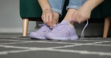 Video unrecognizable cropped woman tie shoelaces on pink sneakers, trying on and testing quality of boots near green armchair. Comfortable orthopedic shoes, body care. Surface low angle view