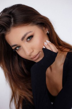 Çekici bir kadın, elleriyle çekici bir şekilde yüzlerine dokunuyor. Kadın stüdyo resmi, yakın plan, beyaz arka plan. Siyah vücut giysisiyle poz veren oldukça kahverengi saçlı bir model. Kadınlık, güzellik.