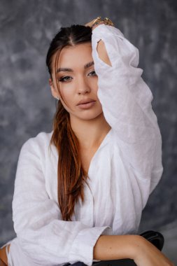 Kolunu kaldırmış, etkileyici bakışları olan zarif seksi bir kadın. Stüdyonun kadın portresi. Güzel kahverengi saçlı kadın beyaz gömlekli poz veriyor. İş görünüşü, temel kıyafetler..