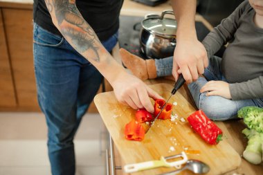 Cropped man with tattoo sleeve cutting pepper food on board near unrecognizable little boy sitting on table. Toddler sitting on kitchen drawer, helping father to cook lunch. Vegetable salad for family