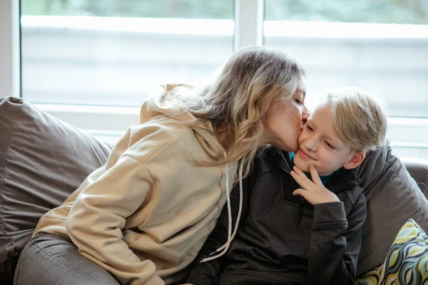 Happy mother and school aged son sitting on sofa near window. Family portrait in cozy home. Woman kiss to cheek little boy, maternal love. Joyful people enjoy comfortable resting. Domestic lifestyle.