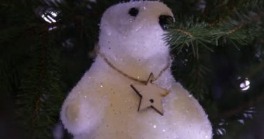 Close-up of white sparkling teddy bear toy with little star on neck hanging on New year Christmas tree decorated with blinking lights garland. New year, Christmas, decoration, celebration, zoom out.