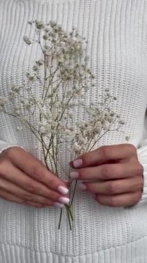 Video vertical format. Womens hands in a knitted cotton sweater with a nude manicure of light shade hold sprig of small white flowers in hands. gentle skin care, natural materials. FullHD footage