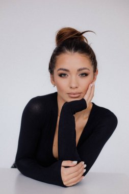 Beautiful woman seductively touching chin with hand. Female studio portrait, white background. Attractive brown haired model with head bun posing in black bodysuit. Fashion, femininity, beauty.