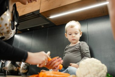 Close up little blond boy sitting on kitchen drawer, looking at vegetable arrangement, while woman peeling carrot and cabbage. Kid development and parent teaching and learning. Low angle view