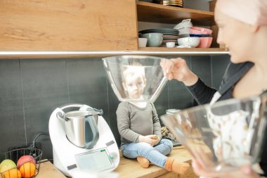 Comic, amazing shot of little cute blond boy sit on wooden kitchen drawer and looking at camera through transparent bowl in smiling woman hands. Cooking with technological kitchenware at home