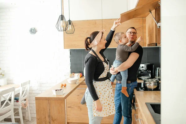 Concentrated, hungry, smiling family of man with boy in hands and woman in apron opening kitchen drawer, thinking what to eat, have lunch. Family cooking and general household chores. Copy space