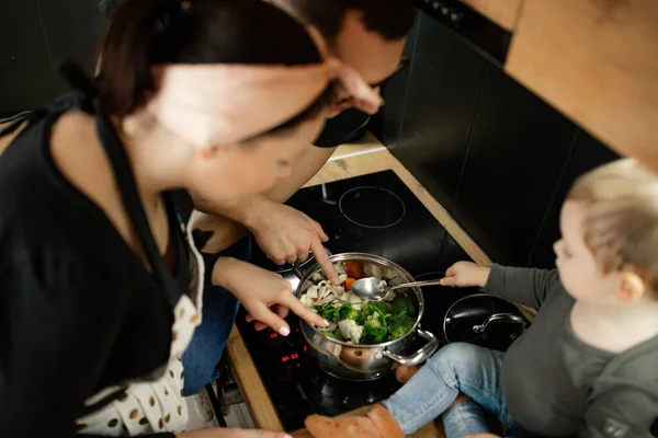 Concentrated family of woman in headband, apron, man and little blond toddler sitting on cooker with vegetable pan in kitchen. Healthy food cooking for whole family. Rearview. High angle view