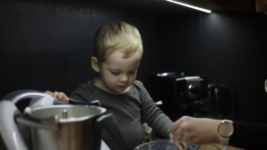 Family of little boy and young woman cooking baking in kitchen at home. Baby toddler sitting on table near kneader cooking machine, playing with flour in bowl, mother holding sons hand with spoon.