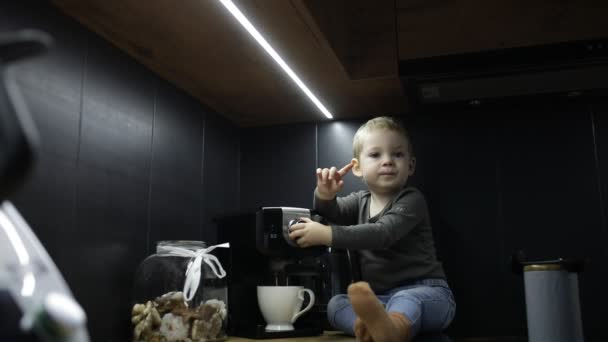 小さな男の子の赤ちゃんは 家庭で黒いキッチンのカップでコーヒーマシンの近くのテーブルの上に座って 電気キッチンのコーヒーメーカーと遊んで 電源を入れます 幼少期の家庭用電化製品 — ストック動画