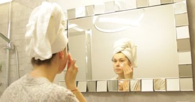 Fresh, nice female with bath towel on head looking at mirror reflection, applying white product moisturizing cream on face. Skin care, face lifting and self massage. Morning beauty routine in bathroom