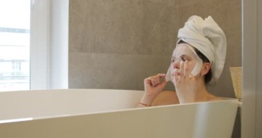 Relaxing young woman sitting in bath in bathroom with hair wrapped in towel, taking off textile mask from face, massaging skin of face with fingers. Cosmetology, skincare, lifting, anti-aging, health.