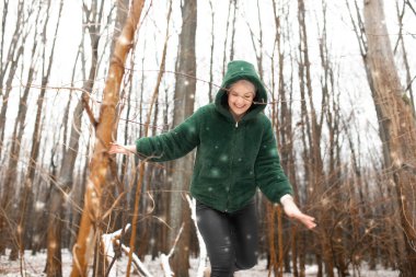 Portrait of funny pretty middle-aged woman wearing green warm jacket, walking running on snow among trees, enjoying weather in park forest in snowy winter, having fun. Winter activities. Snowfall.