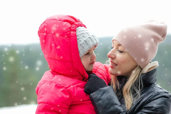 Side view of loving family standing outside in park forest in snowy winter. Middle-aged woman holding little girl, daughter embracing mother, meeting eyes. Motherhood, snow, childhood, relationship.