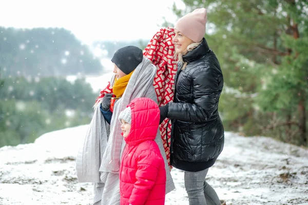 Side view of amazing family standing outside near trees in park forest in snowy winter. Middle-aged woman mother holding red blanket, teenage boy son holding cup near little girl daughter. Snowfall.