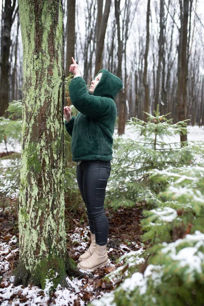 Side view of concentrated middle-aged woman wearing green warm jacket, black jeans, standing outside pointing at green moss on trunk of tree with index finger in park forest in snowy winter. Vertical.