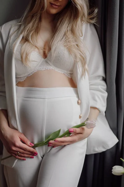 Pregnant blonde with long loose hair stands against gray wall, one hand gently hugs her stomach, in other hand tulip. Maternity clothes. Waiting for miracle. Pregnancy management.