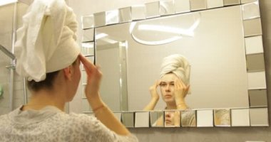 Woman apply moisturizing mask for face, massaging and washing. Camera zooming, closeup. Skincare routine, careful cleaning. Beautiful female wearing towel on head looking in mirror in bathroom.