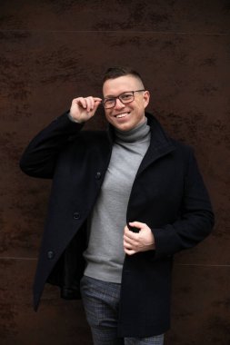 Portrait of successful smiling man in glasses and black cashmere coat standing in front of dark brown wall of office building. Business and entrepreneurship. Career and success. Freelance. 