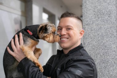 Şirin bir adamın köpeğini kollarına alıp kameraya gülümsediği yakın plan bir görüntü. Evcil hayvanlar. Hayvanların bakımı ve bakımı. Köpeklerle yürümek. Olumlu duygular. Aşk ve arkadaşlık.