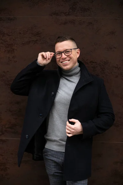 Portrait of successful smiling man in glasses and black cashmere coat standing in front of dark brown wall of office building. Business and entrepreneurship. Career and success. Freelance.