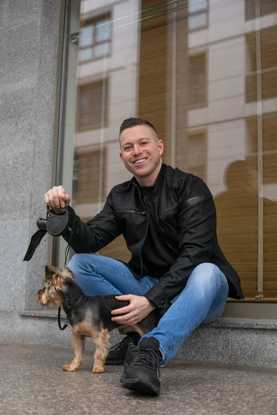 Portrait of young handsome man in jeans and black jacket sitting on street with his dog and looking at camera with smile. Pets. Maintenance and care of animals. Walking with dogs. Positive emotions.