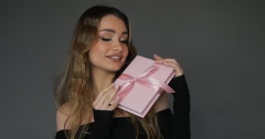 Gülümseyen genç ve çekici bir kadın pembe hediye kutusunu ipek fiyonklu gösteriyor ve farklı yönlere çeviriyor. Stüdyo gri arka plan. Güzellik anlayışı. Hediye çeki. 4k görüntü.